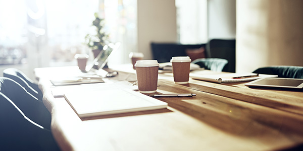 A long wooden conference table set with paper coffee cups, open notebooks, pens, and laptops in a brightly lit meeting room.